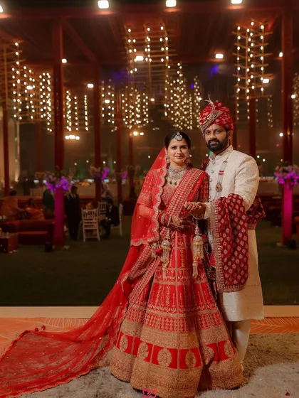 A grand wedding portrait of the couple in their red and white attire, set against the beautifully lit backdrop of their evening reception.