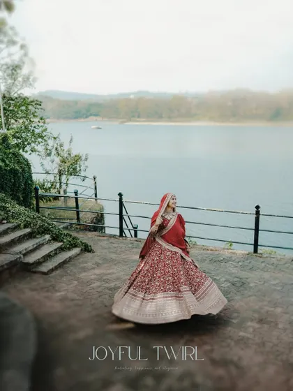 A joyful bride twirls by the water, her red lehenga creating a beautiful splash of color against the serene landscape.