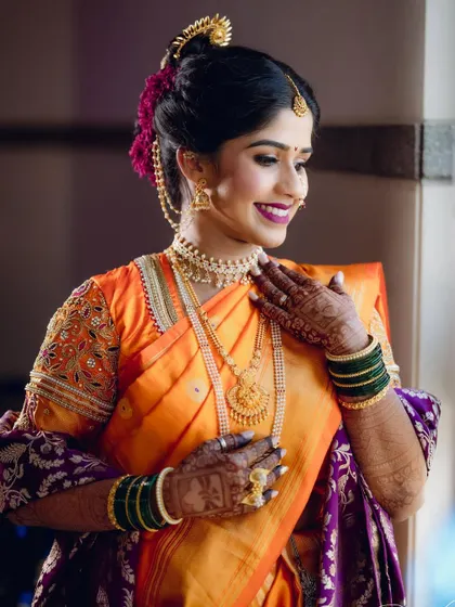 A portrait of a radiant Maharashtrian bride in a bright orange and purple saree, her smile lighting up the room as she gets ready for her big day.