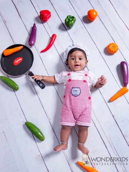 A creative flat-lay photo of a baby dressed as a chef, surrounded by colorful toy vegetables and a frying pan. This top-down perspective is a fun and unique way to do a themed shoot.