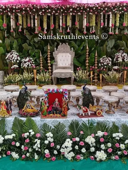 A beautiful baby shower setup with a backdrop of green leaves and hanging pink and white flowers. The empty plates are arranged neatly, ready to be filled with offerings for the ceremony.