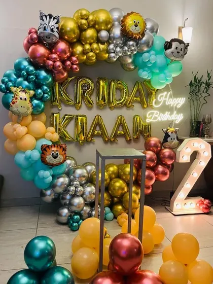 A colorful jungle-themed second birthday. The circular balloon arch is filled with animal foils, the birthday boy's name in foil letters, a neon sign, and a light-up number '2'.