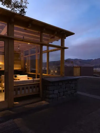 An exterior view of the Postcard hotel at dusk. The building's low profile and use of glass and wood help it sit harmoniously within the fragile Ladakhi landscape, with the mountains in the background.