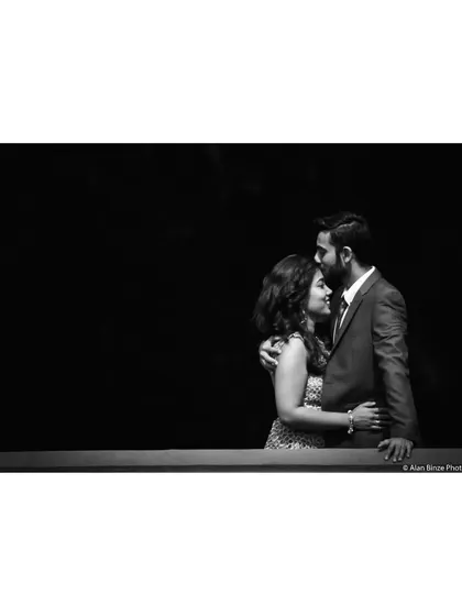An intimate black and white pre-wedding portrait. The high-contrast lighting from the window blinds creates a dramatic and moody effect.