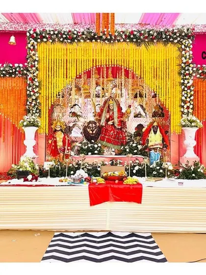 A grand stage setup for a religious function (Jagran). The decor features idols on a beautifully decorated platform with vibrant floral and fabric arrangements.