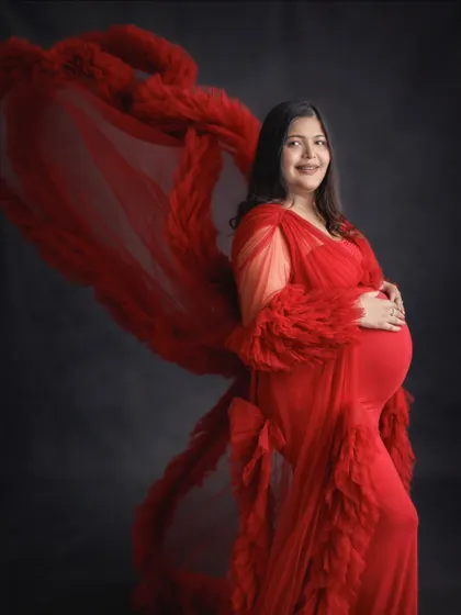 A smiling portrait of a mom-to-be in a red ruffled gown with a flowing train. The dark background makes the red color stand out beautifully.