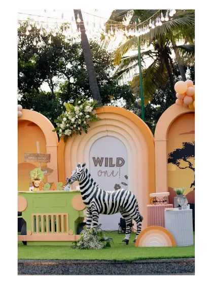 A beautiful sunset-hued 'Wild One' safari party. The setup includes a life-sized zebra prop standing before a custom dessert table, set against a backdrop of layered arches in warm orange and pink tones.