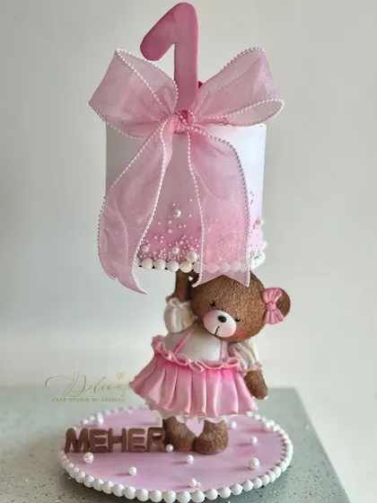 Meet one of our most loved gravity-defying designs. This adorable teddy bear, dressed in a handmade pink frock, holds up a cake for a little one's first birthday. Every detail, from the tiny bow to the delicate pearls, is crafted by hand to make your celebration extra special.