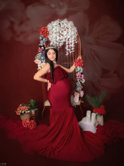 Feeling like royalty in our studio. This mom-to-be wears a crown and a gorgeous red mermaid-style gown, seated in our floral-adorned chair for a truly regal maternity portrait.