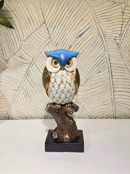 A wise owl perched on a branch, featuring a vibrant blue cap. This colorful resin showpiece is perfect for a study or a child's room.