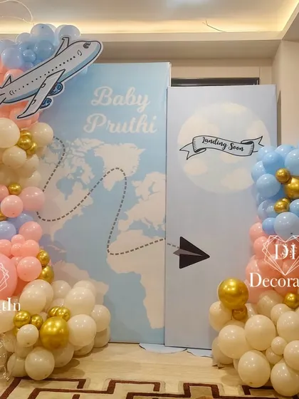 "Baby Pruthi is landing soon." This creative travel-themed baby shower decor features a world map backdrop, an airplane cutout, and a cloud of pink, blue, and gold balloons to celebrate the upcoming arrival.