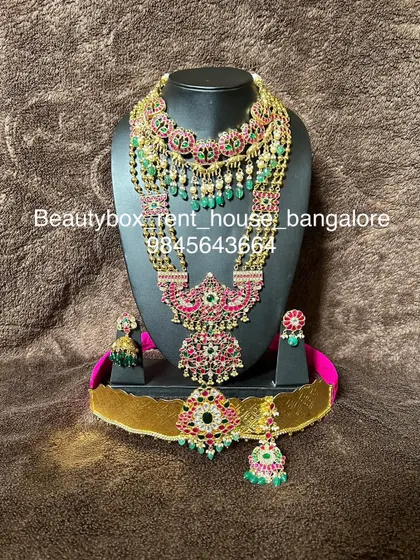 A stunning Jadau-style bridal set with red and green stones and green bead drops. This rental includes multiple necklaces, earrings, maang tikka, and a vaddanam.