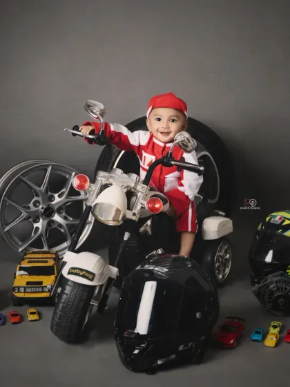 A toddler enjoying his biker-themed photoshoot, sitting on a miniature motorcycle. The set is complete with helmets, wheels, and toy cars for an authentic motor-themed portrait.