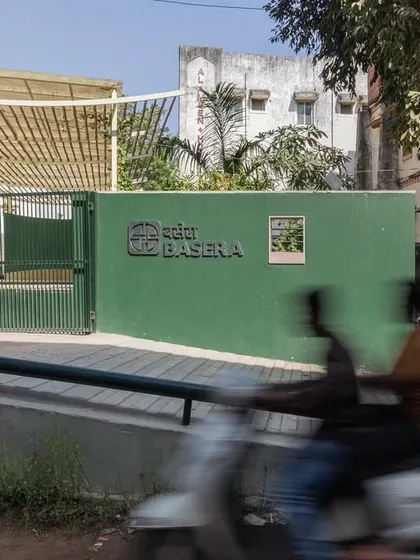 The entrance to Basera Plaza, marked by a simple green wall and clear signage. The motion-blurred scooter in the foreground captures the dynamic urban context in which this quiet community space is nested.