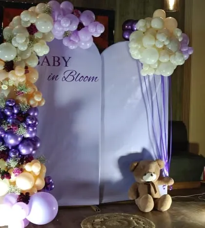 A "Baby in Bloom" baby shower setup in a lovely purple and cream color scheme. The organic balloon arch frames two arched backdrops, with a teddy bear prop adding a cute touch to the arrangement.