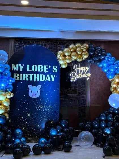 A unique birthday theme with a starry night feel. The dark blue and black backdrop is decorated with a gold and blue balloon arch and a 'Happy Birthday' neon sign.