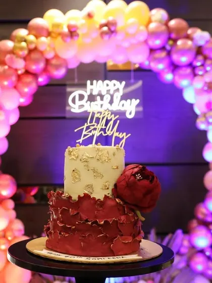 A stunning two-tiered birthday cake with a red floral design, perfectly complementing the colorful balloon arch backdrop.
