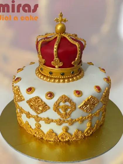 An incredibly ornate crown cake fit for royalty. The white cake is decorated with intricate gold piping and jewels, topped with a stunning red and gold crown.