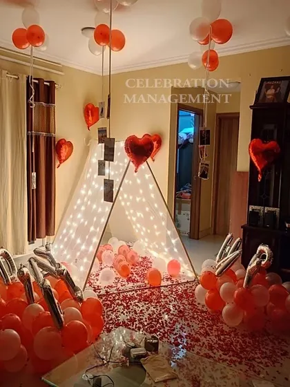 A cozy and personal surprise featuring a lit-up canopy tent in the living room. The floor is covered with rose petals, and photos of special moments hang from the ceiling balloons, making it a walk down memory lane.