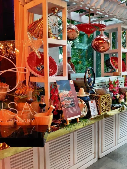 A detailed shot of 'The Malay Satay Street' station. The setup includes thematic props like a conical hat and mortar and pestle, immersing guests in an authentic cultural and culinary experience.