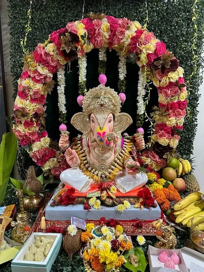A circular floral arch made with multi-colored roses frames the Ganpati idol beautifully. The base is decorated with a variety of fruits and sweets, completing the traditional offering setup.