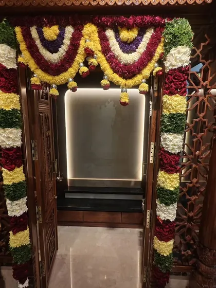A close-up of the intricate floral garlands decorating the entrance of a traditional wooden pooja room for a housewarming ceremony. We use a mix of colorful flowers for a festive look.