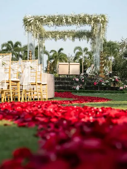 A view down the aisle, covered in a carpet of red rose petals, leading towards the elegant, open-air mandap. This creates a dramatic and romantic path for the bridal entry.