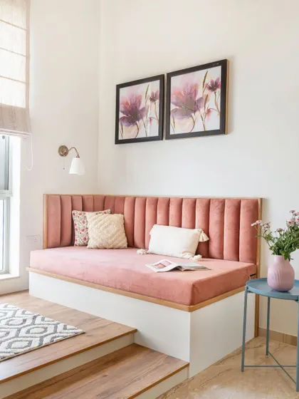 A cozy and inviting reading nook featuring a plush, blush pink platform seat. The corner is elevated on a wooden step and adorned with floral artwork.