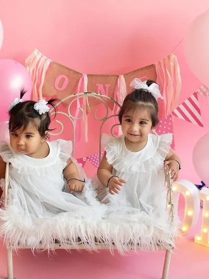 Twinning and winning! The two sisters sitting together on a miniature bed, surrounded by pink balloons, make for an absolutely precious first birthday portrait.