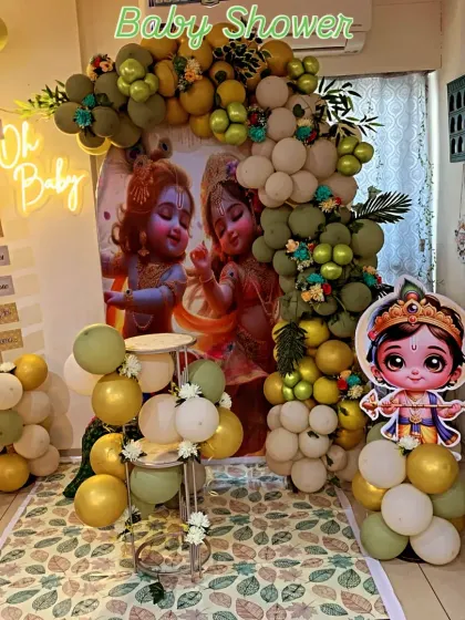 A unique Krishna-themed baby shower. The decoration features a backdrop with cute illustrations of Krishna and Radha, surrounded by a green and gold balloon arch.