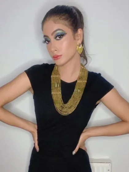 A full-length shot of a glamour fashion look. The makeup features a dramatic blue and green winged eyeshadow, complemented by a bold gold necklace and a sleek high ponytail for a modern, Cleopatra-inspired vibe.