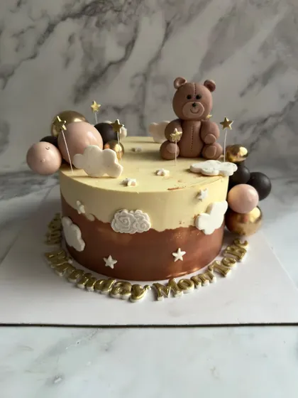 A dreamy teddy bear cake featuring a cute fondant bear sitting amongst clouds, stars, and balloons. The brown and gold color palette gives it a warm and cozy feel.