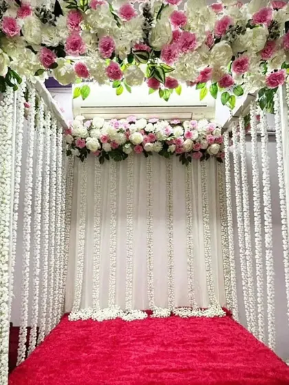 A four-poster mandap design with a canopy of pink and white flowers. The posts are draped with strings of white flowers, creating a beautiful and full look over a red velvet base.
