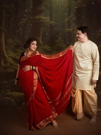 A playful, romantic moment between a couple, set against a mystical forest backdrop. The vibrant red saree stands out, creating a beautiful contrast that highlights the mother-to-be.