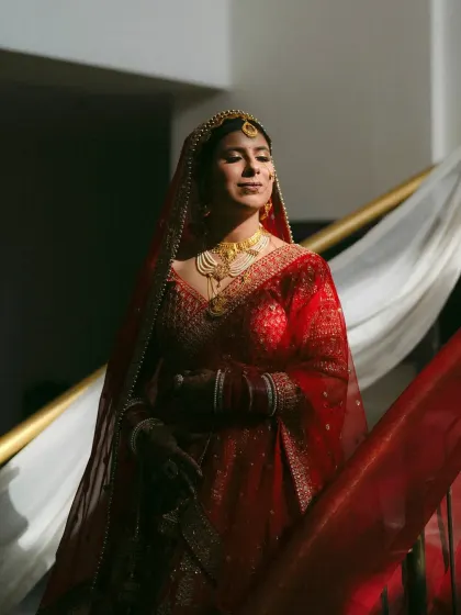 A bride in a rich red lehenga, bathed in dramatic light on a staircase. The image is regal and powerful, highlighting her stunning bridal look.