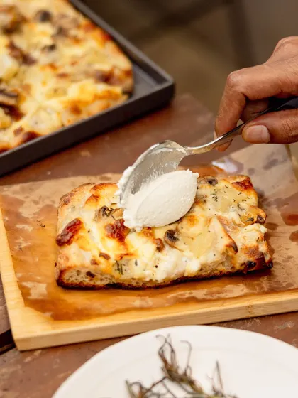 A generous scoop of our house-made ricotta being added to a slice of Bianco Pizza Romana. The creaminess of the ricotta balances the savory roasted potatoes and mushrooms.