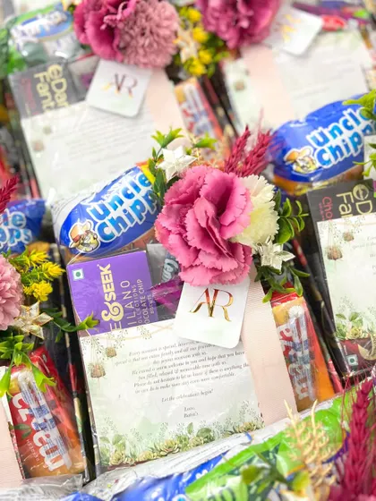 A close-up of our wedding room hampers, filled with snacks like Uncle Chipps and Mazza. Each hamper includes a personalized welcome note and is adorned with a floral touch.