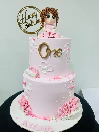 A lovely two-tier pink birthday cake for a first birthday. This cake features delicate white piping, fondant ruffles, and a custom-made doll topper, creating a sweet and personal touch.