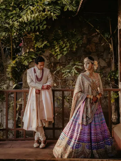 A full-length portrait of a couple standing on a balcony, framed by lush trees. The groom's cream sherwani and the bride's pastel lehenga create a look of understated elegance.