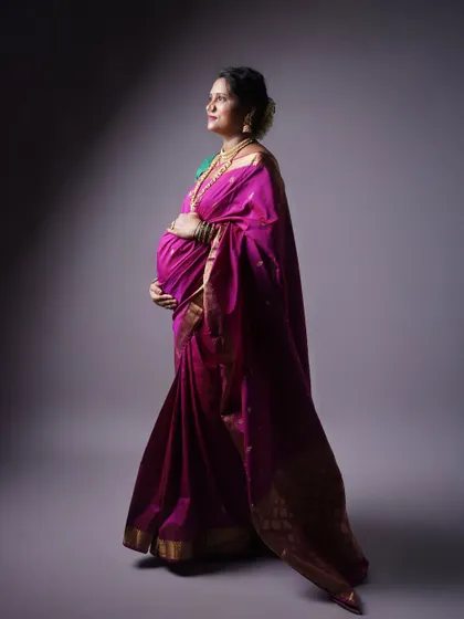 A profile shot of a pregnant woman in a traditional purple saree. The studio lighting is carefully placed to highlight the texture of the fabric and the curve of her belly, creating a classic and respectful portrait.