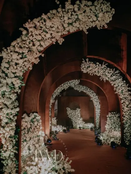A side angle of the Sangeet entrance tunnel, highlighting the contrast between the rustic arches and the delicate white floral arrangements. This is an example of our custom fabrication capabilities.