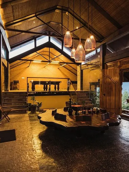 The interior of the Finch Ecofarms guesthouse, featuring a double-height ceiling with exposed wooden beams and a massive, single-piece wooden coffee table. The design emphasizes volume, natural light, and handcrafted details.