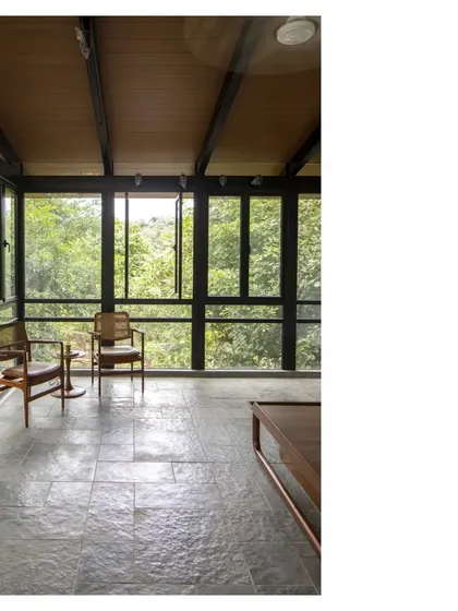 An interior view from the Villa in a Grove, looking out into the dense canopy. The large windows and simple material palette of stone and wood create a calm, protected space that is fully connected to the outdoors.