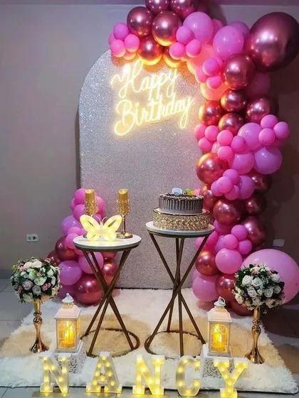 An elegant birthday setup with a glittered arch backdrop and a neon sign. The hot pink balloon garland is paired with decorative lanterns and cake stands.