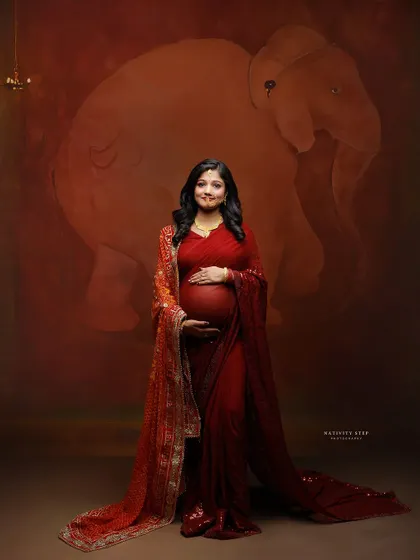 A standing portrait of the mother-to-be in a red saree, her hands gently holding her baby bump. The elephant motif backdrop adds a classic Indian touch.