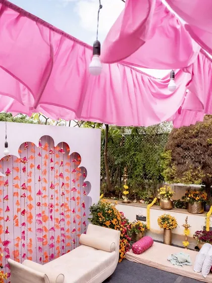 A beautiful and modern Mehendi backdrop set up in the client's home, featuring a scalloped arch with hanging floral tassels and soft pink drapes overhead.
