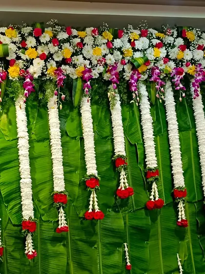 A close-up of a traditional backdrop made with fresh banana leaves. The top is adorned with a colorful mix of flowers and long jasmine garlands, creating a fresh and auspicious look.