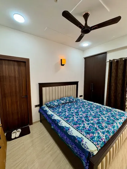 A simple and elegant bedroom design from a 2BHK project. It features a bed with an upholstered headboard, a dark wood wardrobe, and warm wall lighting.