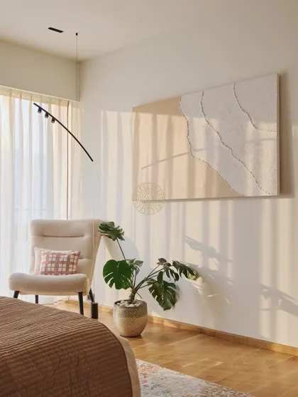 A quiet corner in the master bedroom with a comfortable armchair and a large piece of abstract, textured art. The natural light from the window creates beautiful shadows, enhancing the serene mood.