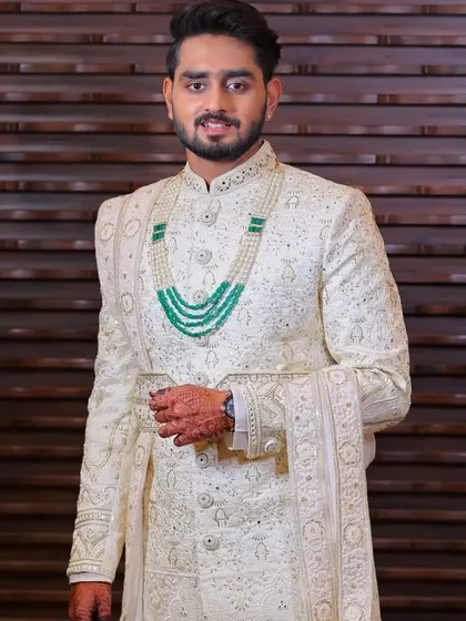 A full-length view of a groom's cream sherwani made from a computerized fabric with katdana work. The look is layered with an anarkali kurta and chudidar, and accessorized with a dupatta and mojari.